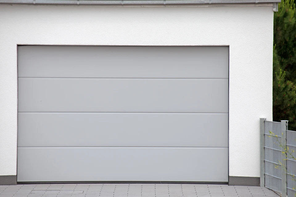 Ext&eacute;rieur r&eacute;sidentiel moderne avec porte de garage sectionnelle grise &agrave; quatre panneaux horizontaux lisses, int&eacute;gr&eacute;e dans fa&ccedil;ade blanche contemporaine