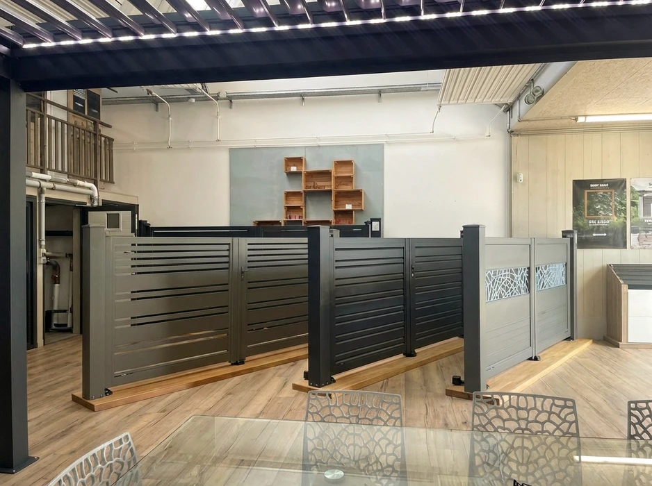 Int&eacute;rieur d'un showroom de menuiserie ext&eacute;rieure bien &eacute;clair&eacute;, avec exposition de plusieurs produits&nbsp;: portails (en aluminium et fer forg&eacute;), pergolas bioclimatiques, stores banne, &eacute;chantillons de fen&ecirc;tres et portes affichant diff&eacute;rentes finitions (couleurs, mat&eacute;riaux). Un couple ou deux clients sont visibles en arri&egrave;re-plan, touchant et examinant les mat&eacute;riaux, discutant avec un vendeur souriantes devant des modules de d&eacute;monstration. L'ambiance est accueillante, le showroom spacieux et bien rang&eacute;, mettant en avant la qualit&eacute; et la vari&eacute;t&eacute; des solutions propos&eacute;es.