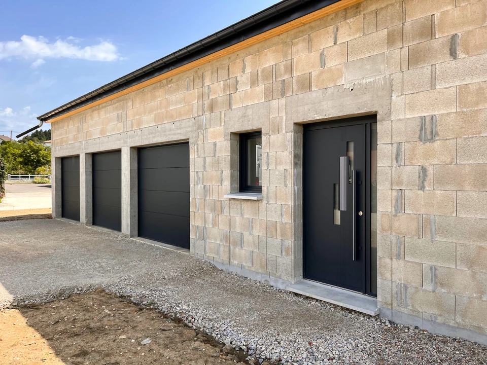 Photo réaliste d'une façade résidentielle moderne montrant l'intégration harmonieuse de plusieurs produits de menuiserie extérieure : fenêtres modernes avec encadrements contemporains, volet roulant intégré, portail d'entrée en aluminium visible au premier plan, le tout illustrant un ensemble cohérent de solutions de menuiserie extérieure sur une même propriété.